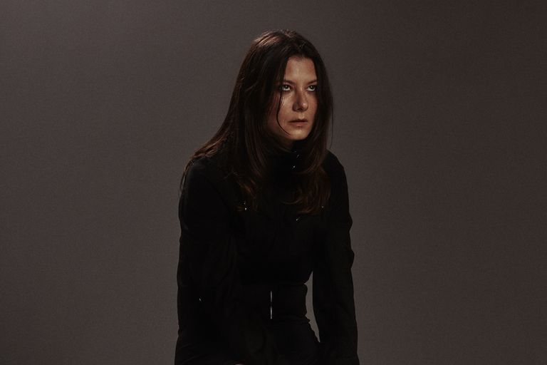 Kate "Babyshakes" Dillon of Full Flower Moon Band in a long-sleeved black outfit, posing against a dark background.